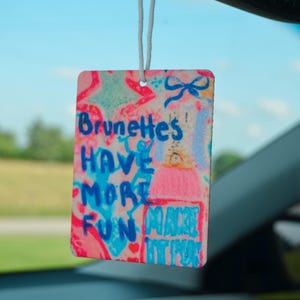 以下が含まれることがあります： 「Brunettes HAVE MORE FUN MAKE IT POP」と青とピンクで書かれた長方形の芳香剤。白い紐で吊り下げられています。背景には青い空と車の内装が写っています。