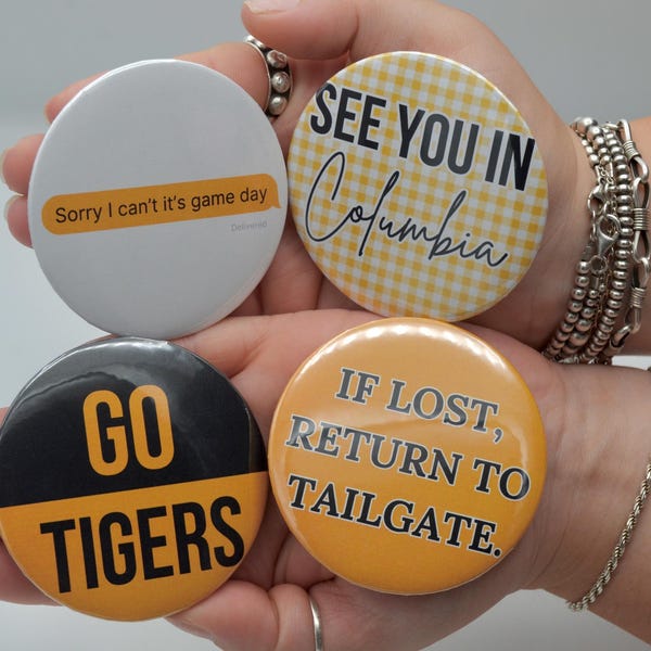 May include: Four round buttons with text. One white button says "Sorry I can't it's game day." Another has "SEE YOU IN Columbia" on a yellow gingham background. A black and yellow button says "GO TIGERS." The last button says "IF LOST, RETURN TO TAILGATE."