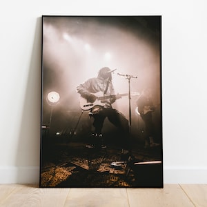May include: A black and white photo of a musician playing a guitar on stage in front of a crowd. The musician is wearing a hooded sweatshirt and jeans. The stage is lit with spotlights and there is smoke in the air.