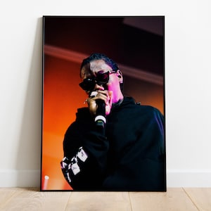 May include: A black and white photo of a person wearing sunglasses and a black hoodie, singing into a microphone on a stage with an orange and purple background.