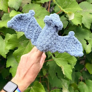 May include: A light blue, handmade crochet bat with outstretched wings. The bat is made of a soft, textured yarn and is held against a backdrop of green leaves. The bat's wings are spread wide, and the body has a rounded shape.