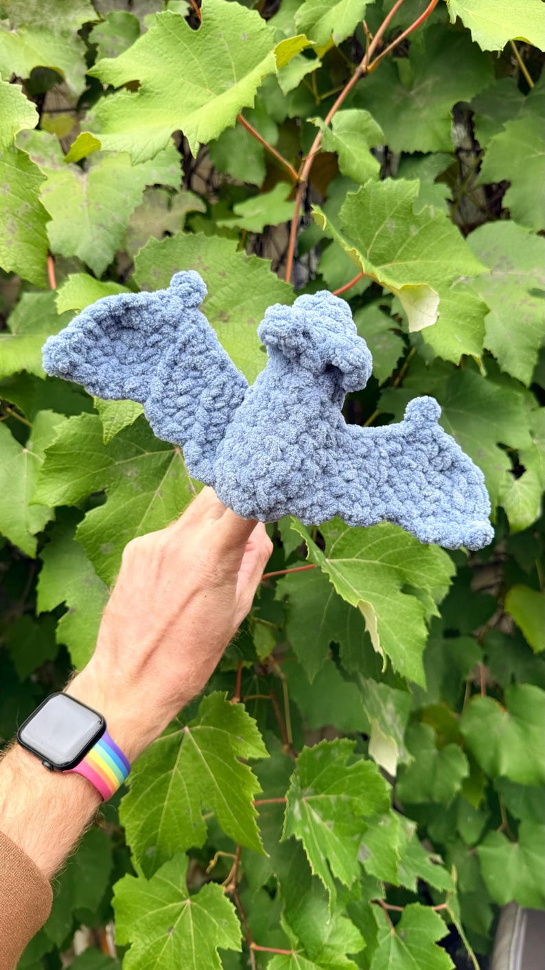 May include: A light blue, handmade crochet bat with outstretched wings. The bat is made of a soft, textured yarn and is held against a backdrop of green leaves. The bat's wings are spread wide, and the body has a rounded shape.