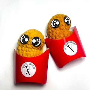 May include: Two crocheted fried chicken nuggets with googly eyes in red cardboard containers. The containers have a white circle with the text "The Crochet Carpenter" and a black hammer and nail.