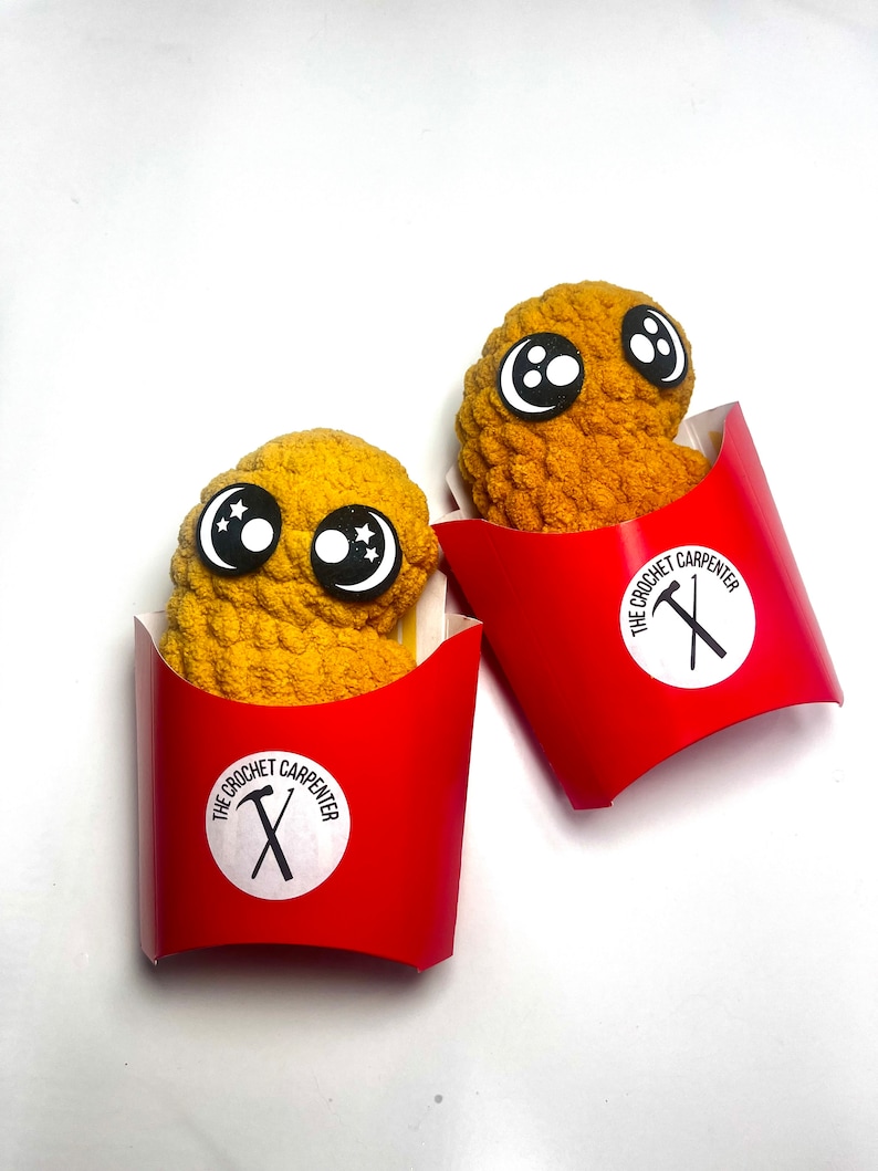 May include: Two crocheted fried chicken nuggets with googly eyes in red cardboard containers. The containers have a white circle with the text "The Crochet Carpenter" and a black hammer and nail.