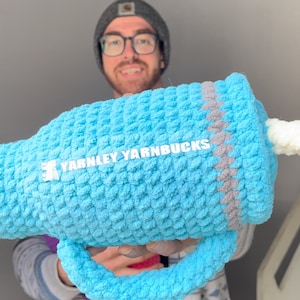 Op de afbeelding: Een groot, lichtblauw gehaakt item met een handvat, met de tekst "YARNLEY YARNBUCKS" in het wit. Het item heeft een getextureerd oppervlak en is geaccentueerd met grijs. Het algemene thema is handgemaakte ambachten.