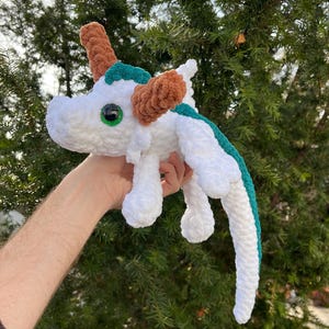 May include: White crocheted dragon plush toy with green and brown accents. The dragon has large green eyes and a long tail.