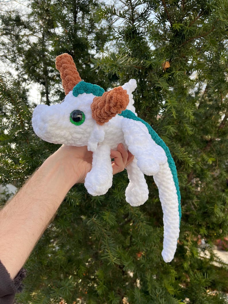 May include: White crocheted dragon plush toy with green and brown accents. The dragon has large green eyes and a long tail.