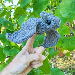 May include: A handmade, gray crocheted dragon with black eyes and outstretched wings. The dragon is held up against a backdrop of green leaves and branches, showcasing its textured, soft appearance.