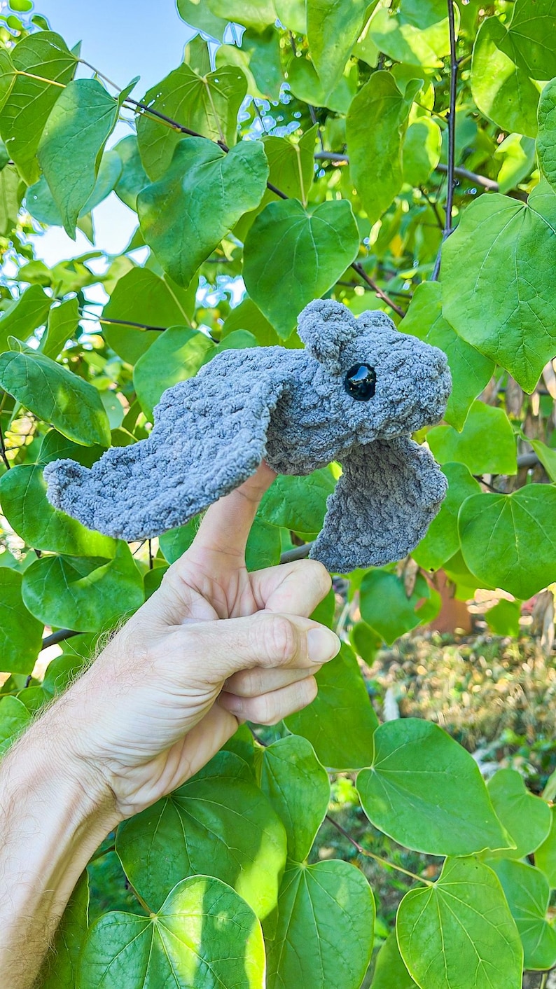 May include: A handmade, gray crocheted dragon with black eyes and outstretched wings. The dragon is held up against a backdrop of green leaves and branches, showcasing its textured, soft appearance.