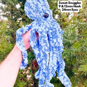 May include: A blue crocheted octopus with white flecks. The octopus has large black eyes and is held by a hand. The text "Sweet Snuggles 9 & 12mm Hook 24mm Eyes" is in the upper right corner of the image.