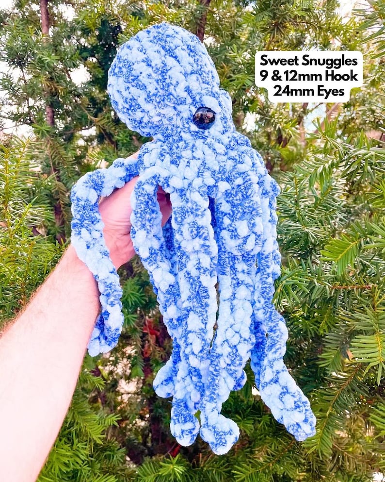 May include: A blue crocheted octopus with white flecks. The octopus has large black eyes and is held by a hand. The text "Sweet Snuggles 9 & 12mm Hook 24mm Eyes" is in the upper right corner of the image.