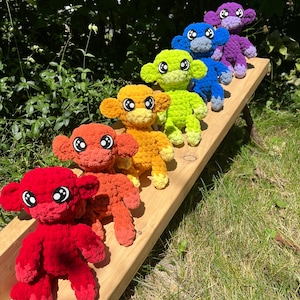 May include: A row of seven crocheted monkey toys in rainbow colors, sitting on a wooden bench. The monkeys are arranged from red to purple, with each monkey facing forward.