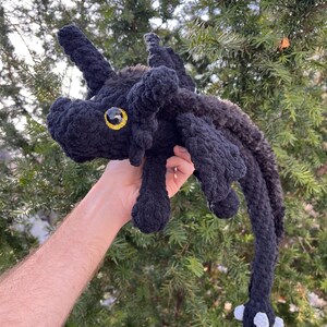 May include: A black crocheted plush dragon with a yellow eye and white feet. The dragon is being held in a person's hand.