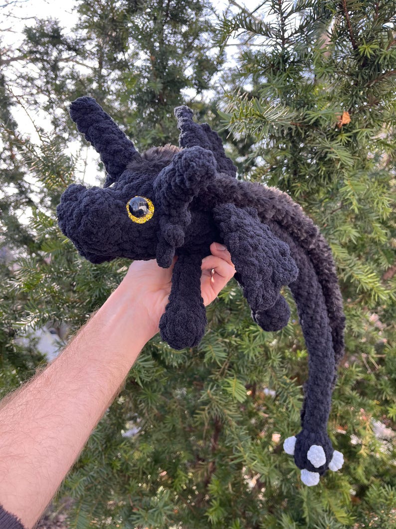 May include: A black crocheted plush dragon with a yellow eye and white feet. The dragon is being held in a person's hand.