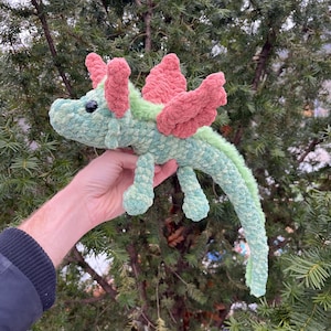 May include: A green and pink crocheted dragon toy with a long tail and wings. The dragon has a small, black eye.