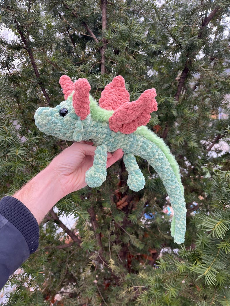 May include: A green and pink crocheted dragon toy with a long tail and wings. The dragon has a small, black eye.