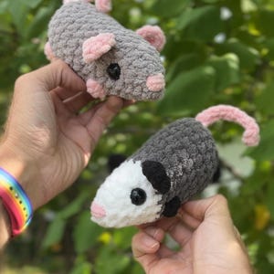 May include: Two handmade crochet possums. One is gray with pink ears and tail, the other is gray and white with black ears. Both have black eyes. The possums are held up against a background of green leaves.