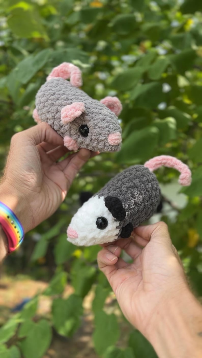 May include: Two handmade crochet possums. One is gray with pink ears and tail, the other is gray and white with black ears. Both have black eyes. The possums are held up against a background of green leaves.