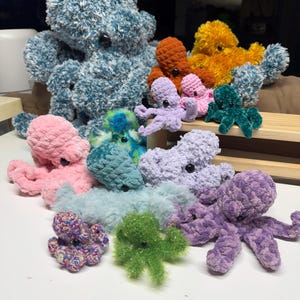 May include: A selection of handmade crochet octopus plush toys in various sizes and colours. The plushies range from large teal and orange to small pink and purple, with button eyes. The soft toys are arranged on a white surface.