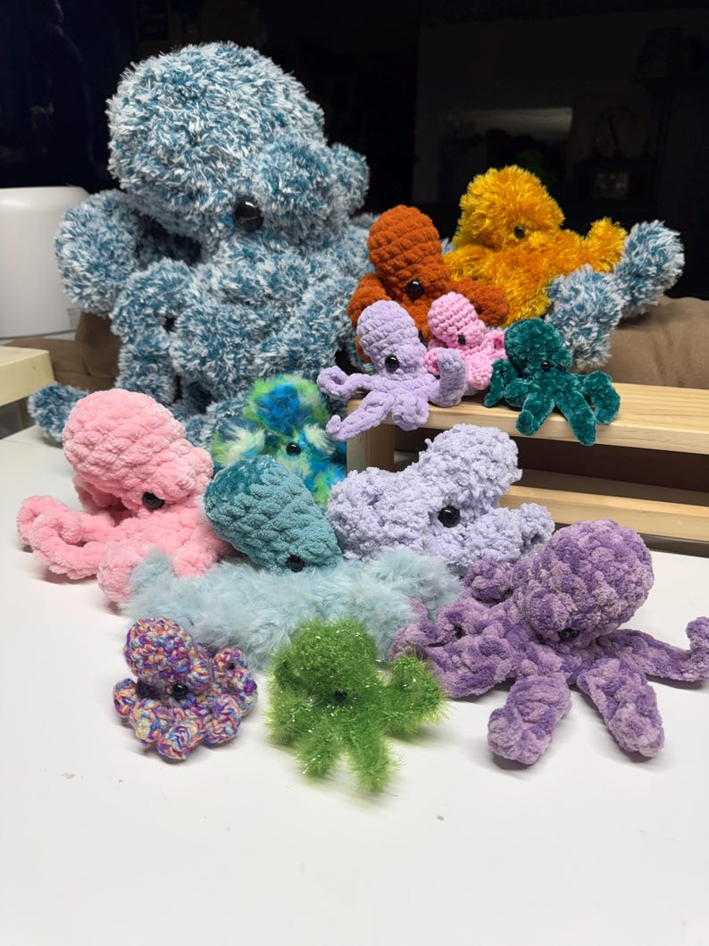 May include: A selection of handmade crochet octopus plush toys in various sizes and colours. The plushies range from large teal and orange to small pink and purple, with button eyes. The soft toys are arranged on a white surface.