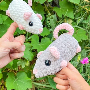 May include: Two crocheted toy rats, one white and one gray, each with pink ears, feet, and tails. The rats have black button eyes. The toys are held up against a background of green foliage.