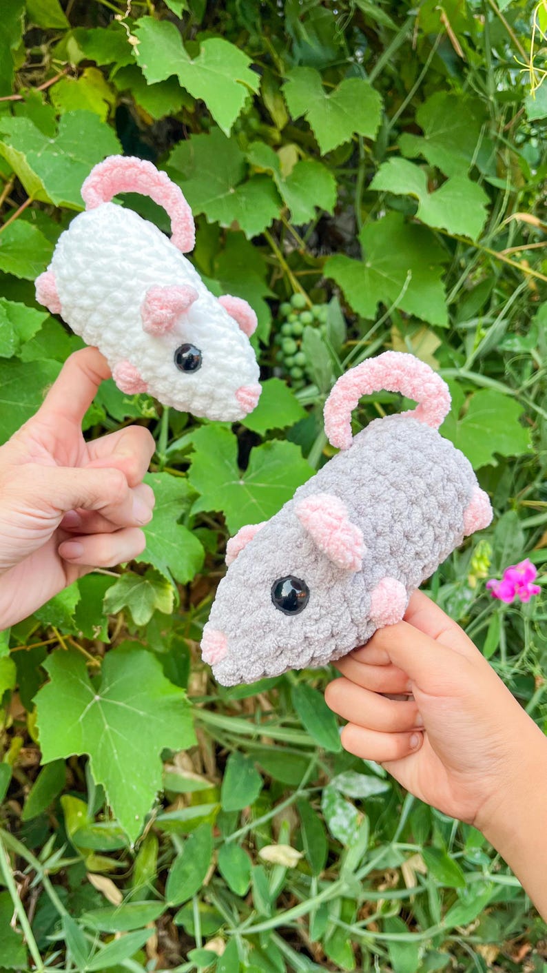 May include: Two crocheted toy rats, one white and one gray, each with pink ears, feet, and tails. The rats have black button eyes. The toys are held up against a background of green foliage.