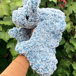 May include: A plush, blue and white stuffed animal shaped like a hippopotamus. The toy has a fluffy texture and a small black eye. The background features green foliage.