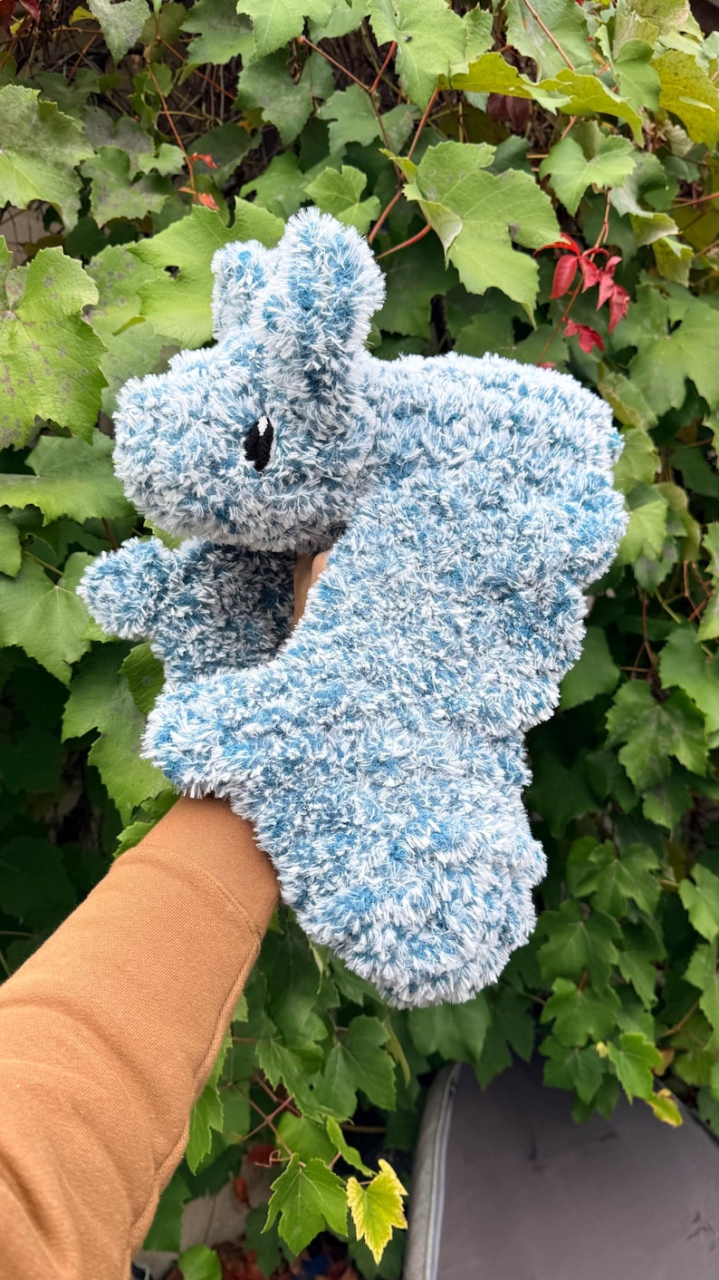 May include: A plush, blue and white stuffed animal shaped like a hippopotamus. The toy has a fluffy texture and a small black eye. The background features green foliage.