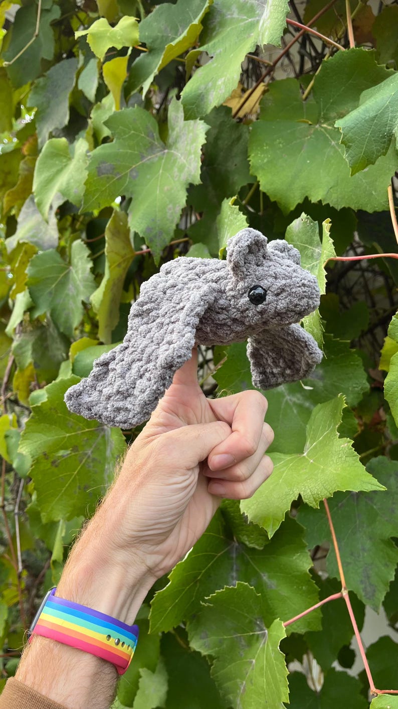 May include: A gray crocheted bat with black eyes and outstretched wings. The bat is held up against a backdrop of green leaves. The bat's wings are spread wide, and its body is made of a soft, textured yarn.