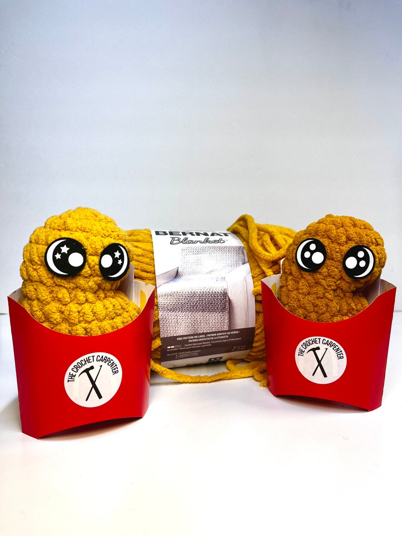 May include: Two yellow crocheted amigurumi toys shaped like french fries in red and white boxes with the text "The Crochet Carpenter" and a crossed hammer and saw. A ball of yellow yarn with a label that reads "Bernat Blanket" is in the background.
