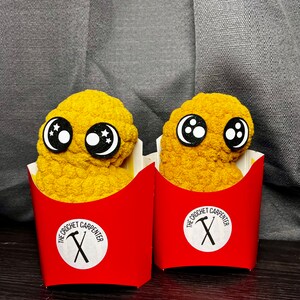 May include: Two yellow crocheted chicken nugget toys with black eyes and white pupils. Each nugget is in a red cardboard container with the text "The Crochet Carpenter" and a crossed hammer and saw logo.
