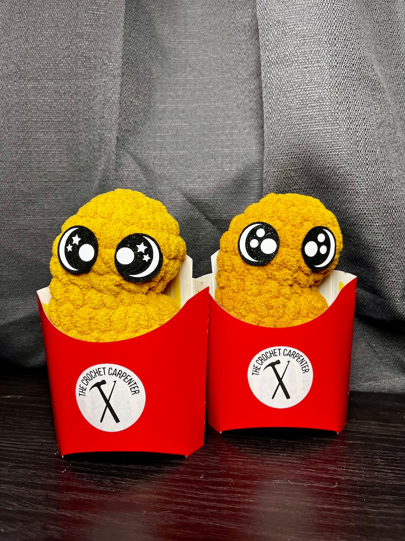 May include: Two yellow crocheted chicken nugget toys with black eyes and white pupils. Each nugget is in a red cardboard container with the text "The Crochet Carpenter" and a crossed hammer and saw logo.