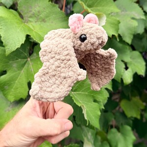 May include: A handmade, plush bat toy in shades of tan and pink. The bat has large, textured wings and small pink ears. The toy is held in front of a backdrop of green leaves.