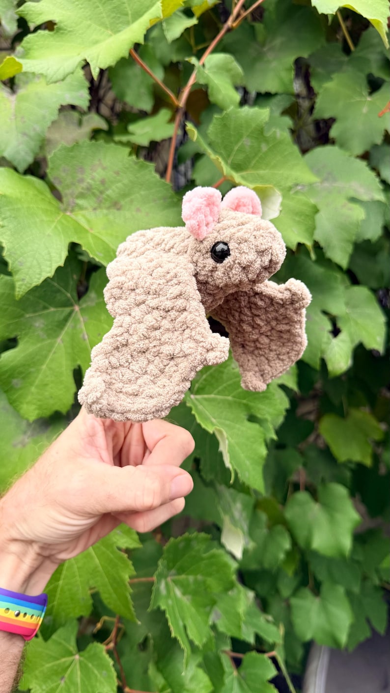 May include: A handmade, plush bat toy in shades of tan and pink. The bat has large, textured wings and small pink ears. The toy is held in front of a backdrop of green leaves.