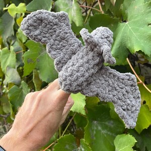 May include: A gray, crocheted bat with outstretched wings. The handmade bat is made of a soft, textured yarn. The bat is displayed against a backdrop of green leaves.