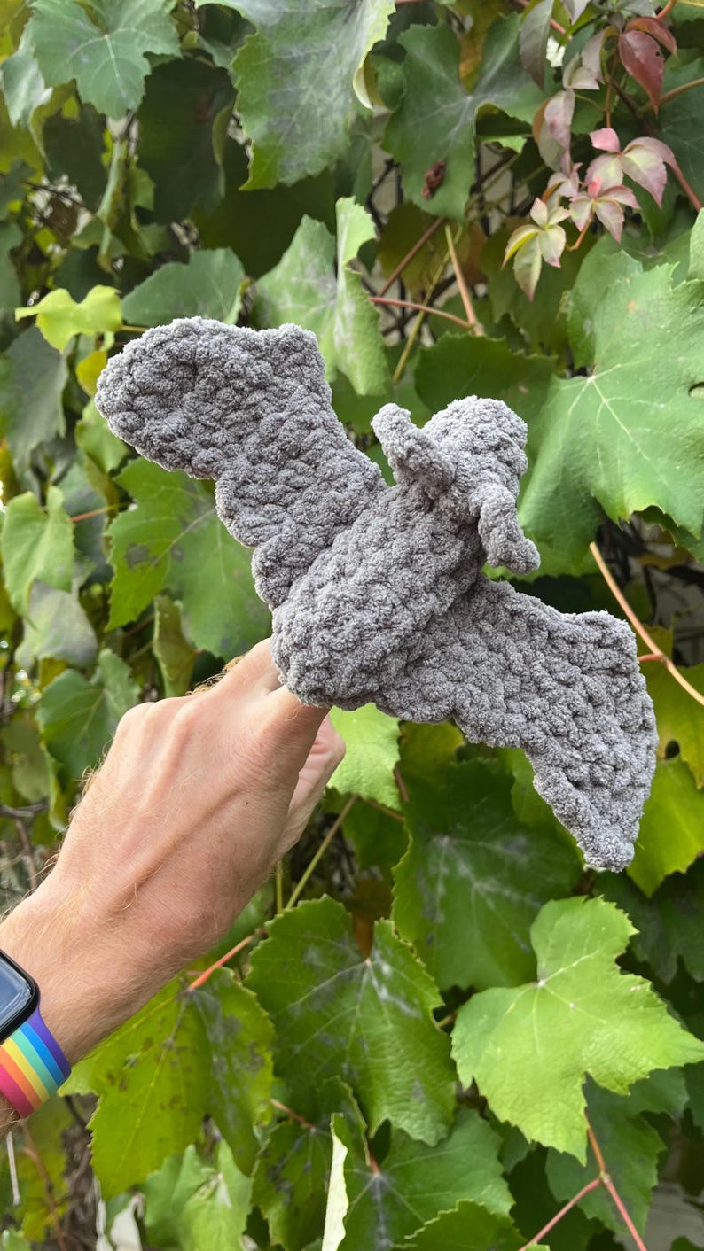 May include: A gray, crocheted bat with outstretched wings. The handmade bat is made of a soft, textured yarn. The bat is displayed against a backdrop of green leaves.