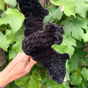 May include: A handmade, black, plush bat, held in front of a background of green leaves. The bat is made of a soft, textured material, with detailed wings and body. The bat is a Halloween or gothic-themed item.