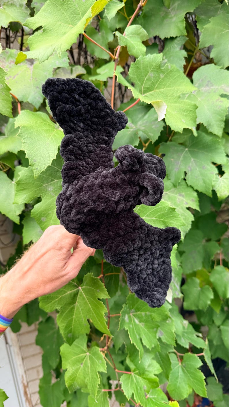 May include: A handmade, black, plush bat, held in front of a background of green leaves. The bat is made of a soft, textured material, with detailed wings and body. The bat is a Halloween or gothic-themed item.
