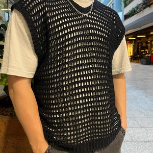 Peut inclure: Un gilet noir en crochet à mailles ajourées, porté sur un t-shirt blanc. Le gilet sans manches présente un motif texturé. Un collier en chaîne argentée est visible. La personne porte un jean gris foncé. L'arrière-plan montre un centre commercial extérieur moderne.