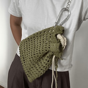 May include: An olive green crocheted bag with a drawstring closure and a silver chain strap. The bag is attached to a large silver carabiner clip. The bag's open weave design reveals a cream-coloured lining.