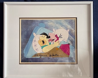 May include: A framed cartoon cel featuring Fred Flintstone and Barney Rubble in bed, signed by Bill Hanna and Joe Barbera. The cel is set against a blue and white background.