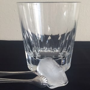 May include: A clear glass with a cut design sits on a black surface. A single ice cube is held by silver tongs.