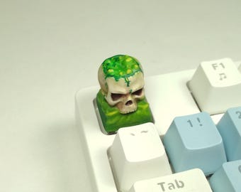 Puede incluir: Una tecla personalizada con un diseño de calavera y detalles verdes, colocada en un teclado mecánico blanco y azul claro. La calavera tiene un aspecto desgastado, y la tecla se asienta sobre una base verde.