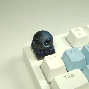 Può includere: Un copritasto personalizzato a forma di Morte Nera, in nero e blu, su una tastiera meccanica bianca e blu. Il copritasto mostra i dettagli della superficie. La tastiera ha tasti bianchi e azzurri.