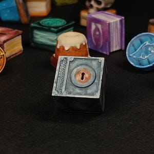 May include: A collection of miniature fantasy-themed objects, including a skull, books, coins, and bottles. The objects are painted in various colors, such as red, blue, gold, and green. A small, gray box with a keyhole is in the foreground.