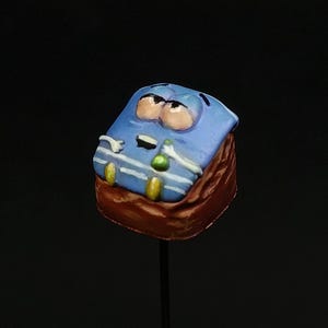 May include: A whimsical, hand-painted keycap featuring a cartoonish face with large eyes and a sad expression. The keycap has a blue top with white stripes and a brown base, mounted on a black stem. The design includes yellow and green accents.