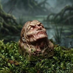 May include: A sculpted figurine of a human head with long hair, pale skin, and a weathered, grimacing expression. The base of the sculpture features textured, moss-like green details, and the overall appearance is aged and distressed in tone.