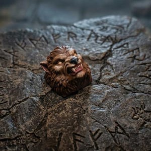 May include: A brown and tan werewolf head figurine with bared teeth and pointed ears, resting on a dark gray stone slab featuring carved runic symbols and etched animal figures across its textured, cracked surface.