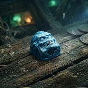 May include: A blue mechanical keyboard keycap sculpted in the shape of a weathered, skeletal face with visible teeth and deep-set eyes. The keycap features a detailed, textured surface and is positioned on a dark, wooden plank surface.