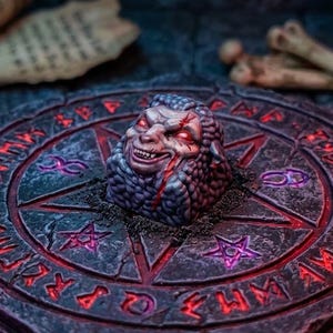 May include: A sculpted artisan keycap featuring a detailed, textured creature head with glowing red eyes, centered on a circular stone-like base engraved with a star and runic symbols illuminated by a vibrant red light.